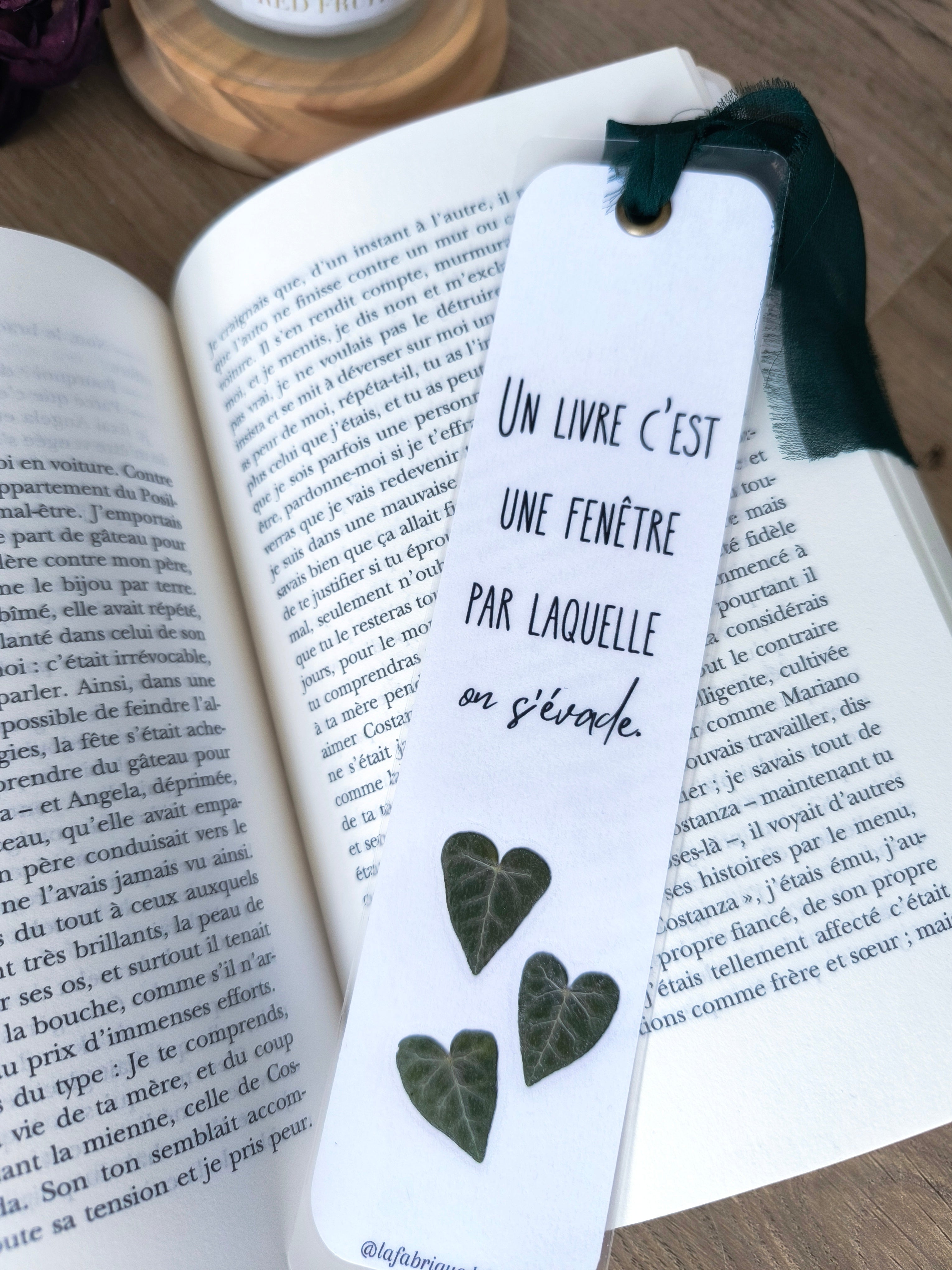 Marque-page en papier avec feuilles vertes en forme de cœur et citation 'Un livre c'est une fenêtre par laquelle on s'évade', posé sur un livre ouvert.