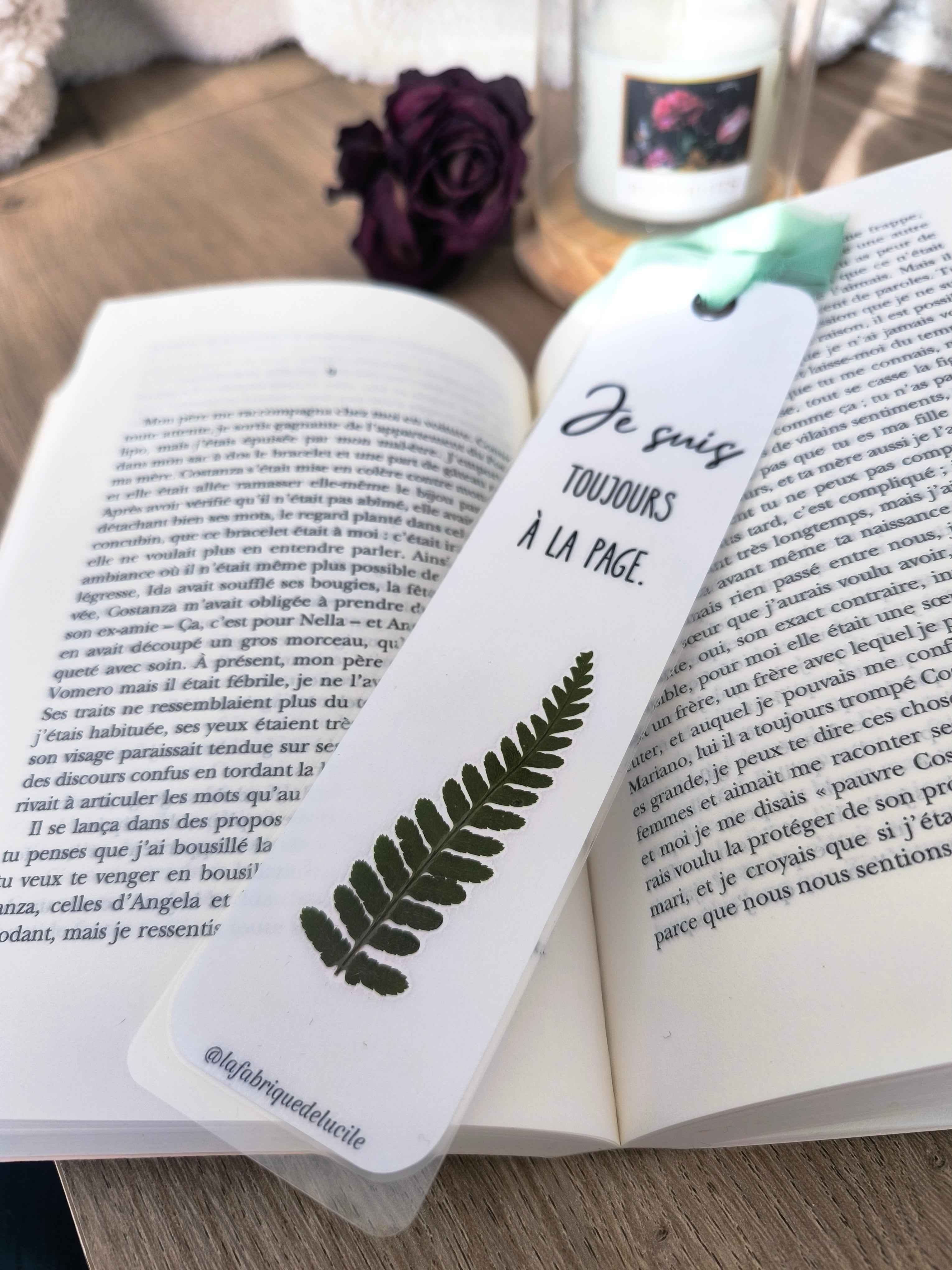 Marque-page en papier blanc avec feuille de fougère séchée et texte 'Je suis toujours à la page', posé sur livre ouvert.